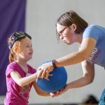 Adult instructor handing a blue goalball to a young redhead girl.