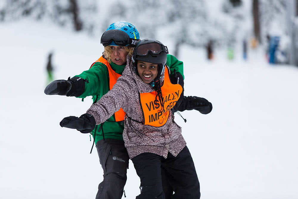 A young athlete being taught to snowboard by a volunteer.