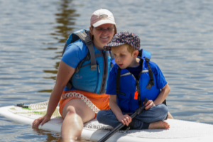 Female guide sitting on a paddleboard with a young boy athlete sitting in front looking out at the water.