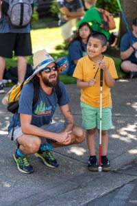 Camp counselor squatting down to talk to a young camper holding a cane.