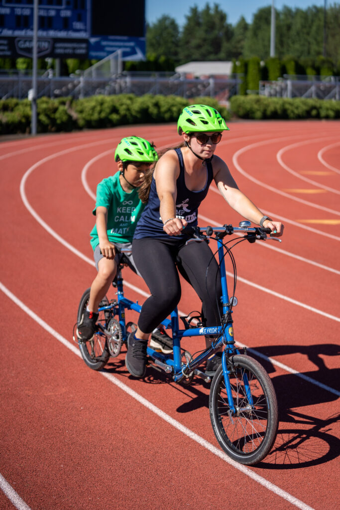 A female counselor is riding tandem with a young boy on the back. Both are wearing fluorescent green helmets.