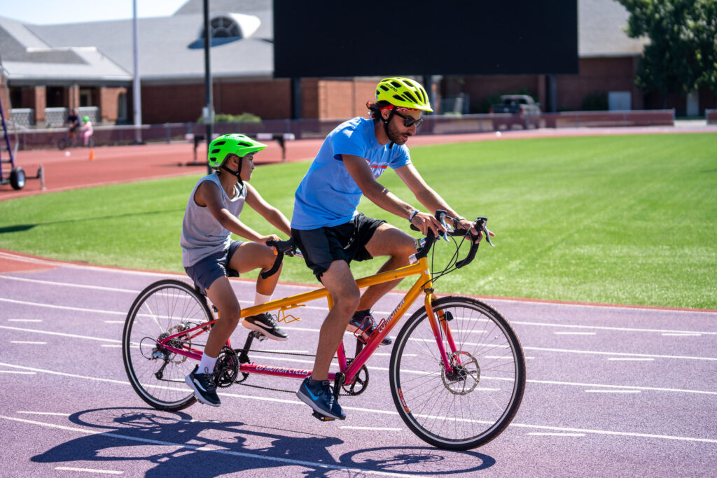 Camp Spark staff member riding on a tandem bicycle with a young camper.