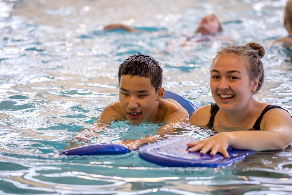 Female volunteer swimming beside a young boy and they're both holding on to kickboards.