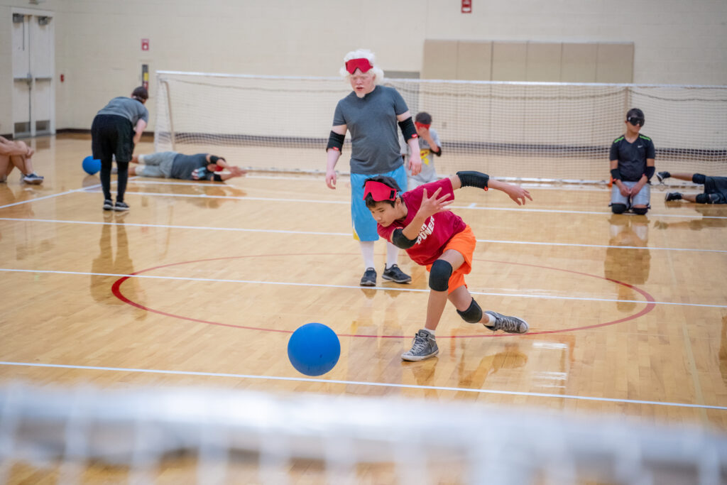 Young boy throwing a goalball as an adult teammate looks on from behind.