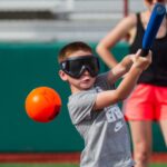Young boy wearing eyeshades swinging a bat towards an orange ball.