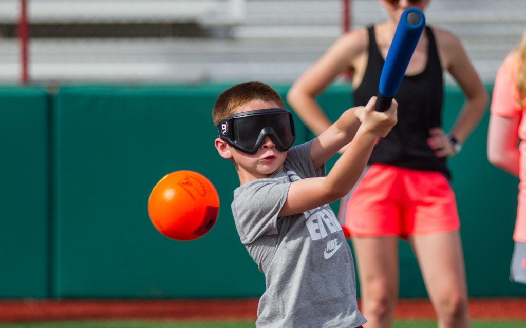 Young boy wearing eyeshades swinging a bat towards an orange ball.