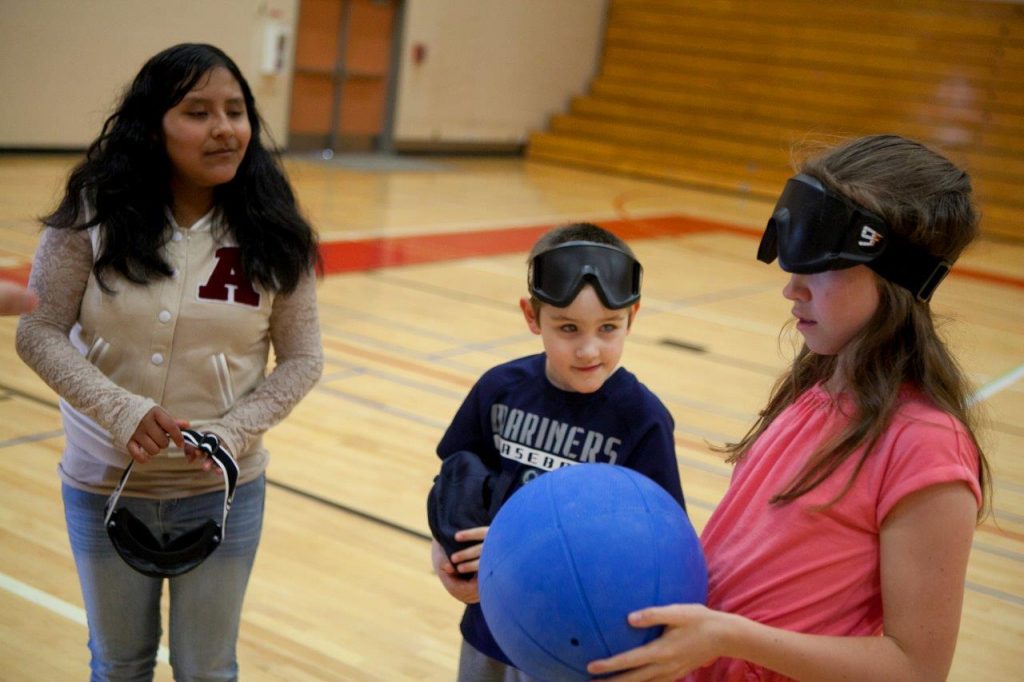 A young girl wearing eye shades is in a gymnasium holding a goal ball while two other students look on.