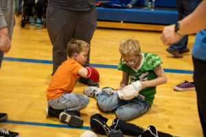 Two young boys sitting on a gym floor putting on protective padding.