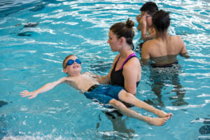 Young Kaleb floating in a pool wearing swim goggles is held up by a Camp Spark counselor.