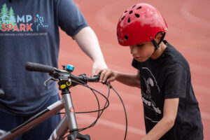 Young athlete wearing a red bike helmet looks over his bike for safety while a counselors arms hold the bike up.