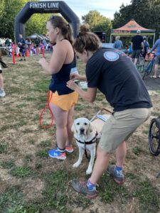 NWABA team member helping participant, Kirsten French by attaching her race bib to the back of her shirt as guide dog, Knightley looks on.