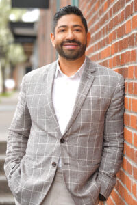 Angel Reyes wearing a white button down shirt and grey patterned suit coat leaning against a brick wall.