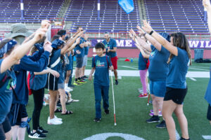 Young boy wearing a Camp Spark tshirt and holding a white cane walking through a human tunnel with everyone holding their hands in the air.