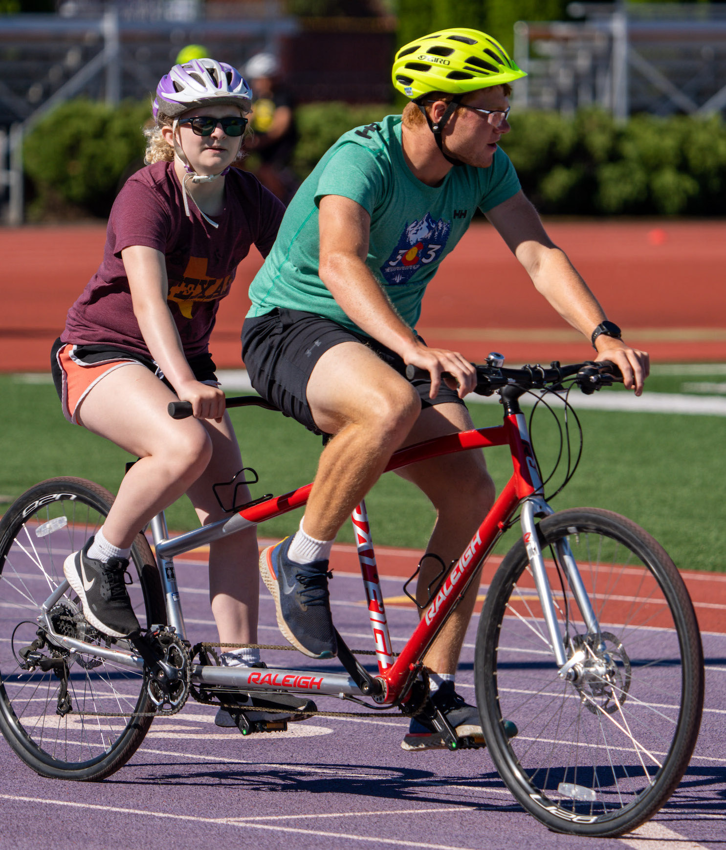 Young athlete, Alaina, riding on the back of a red tandem bicycle with her guide in the front seat. Alaina is wearing a burgundy tshirt, orange shorts and a white helmet.