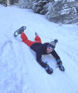 NWABA staff member, Matt sliding down a snowbank wearing snowshoes and a knit cap, looking up at the camera smiling.