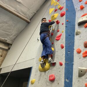 Image of athlete, Umberto, hanging onto an indoor rock climbing wall while harnessed and looking down at the camera.