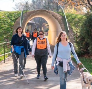 Two athletes and their guide are walking through a park tunnel. Left to right: adult woman in navy shirt holding a cane, sighted guide is wearing an orange vest with "guide" across the front, young woman in a light blue t-shirt is holding the harness of her yellow lab guide dog.