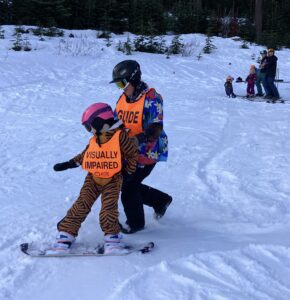 Young athlete in a tiger suit and pink helmet is learning to snowboard with an instructor guide standing behind her.