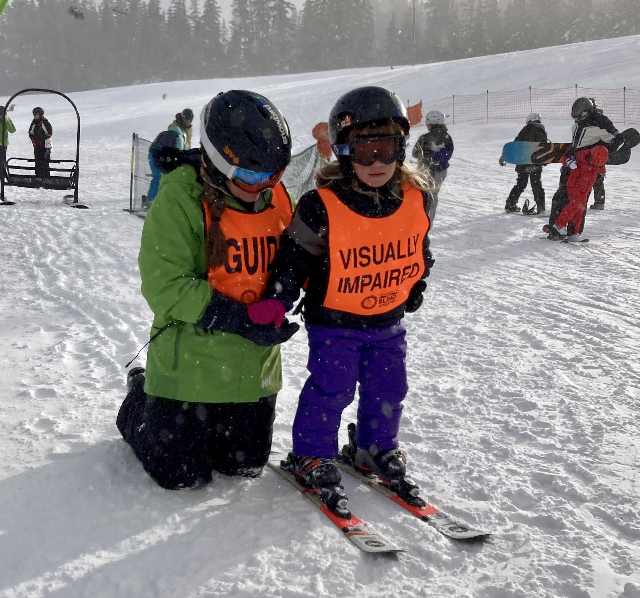 Young athlete on skiis wearing purple pants, a black helmet, goggles and an orange safety vest. An adult guide is behind the athlete on her knees holding her hand for stability. The guide is wearing a green ski jacket, helmet and goggles. Other skiers and snowboarders are walking behind them.