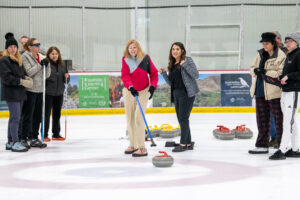 A group of athletes and volunteers surround a female athlete and look on while she sweeps a curling stone across the ice.