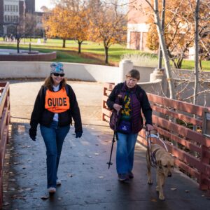 An athlete and guide are walking across a footbridge on a fall day. The athlete is holding her guide dog while the guide is walking alongside her.