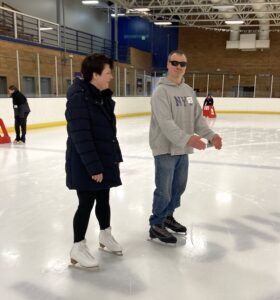 A male NWABA athlete is wearing jeans and a grey hooded sweatshirt while gliding on ice skates. A female guide wearing a black coat follows alongside giving guidance.