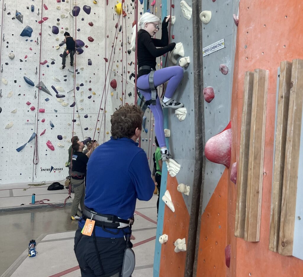 Young blonde athlete is climbing an indoor rock wall wearing a harness as a volunteer is below holding the ropes for safety