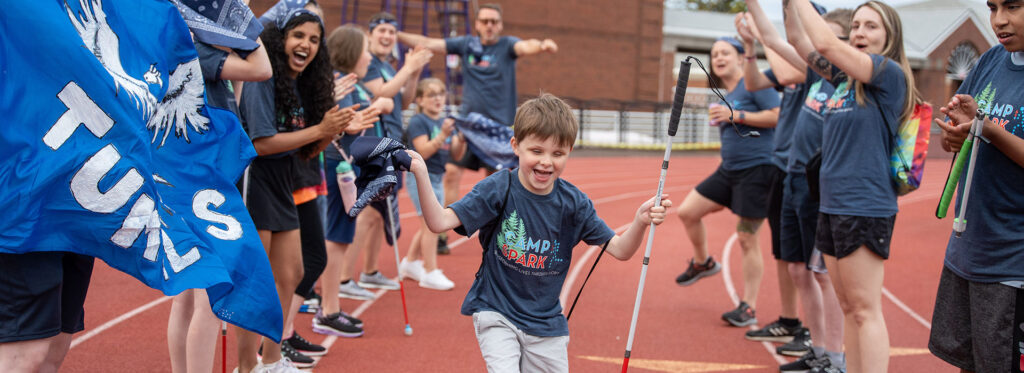 A young boy running with a white cane through a tunnel of cheering campers with their arms up. Everyone is smiling and cheering in Camp Spark shirts