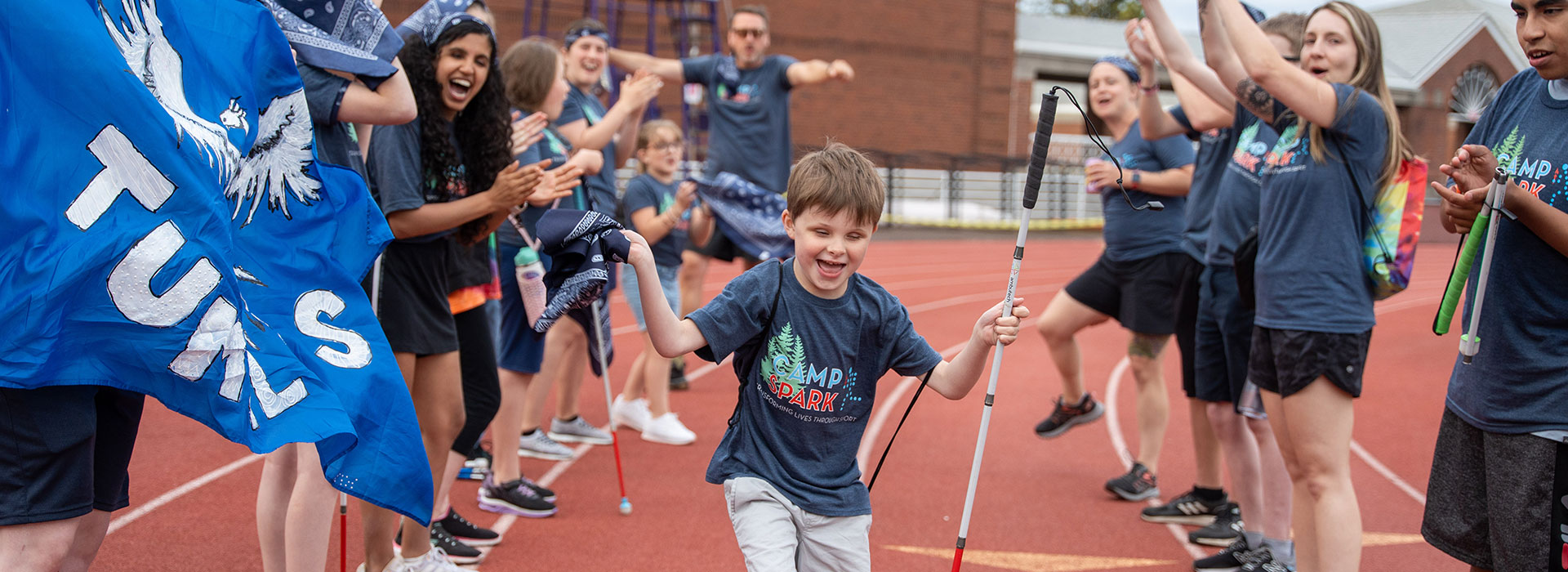 A young boy running with a white cane through a tunnel of cheering campers with their arms up. Everyone is smiling and cheering in Camp Spark shirts