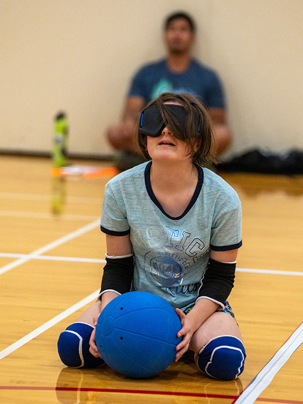 young girl kneeling ina gym holding a goalball and wearing eyeshades