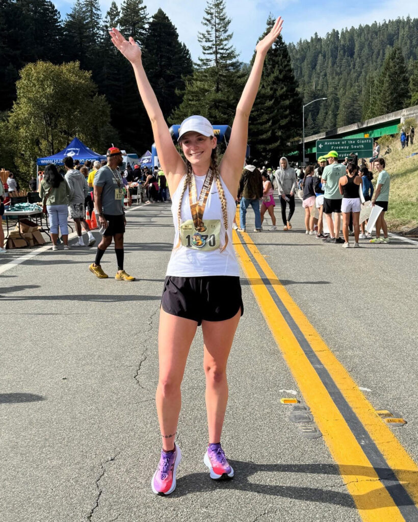 Rachelle Standing in the road with other runners after a race. The sun is shining, she is smiling, and her hands are up in celebration for finishing a race