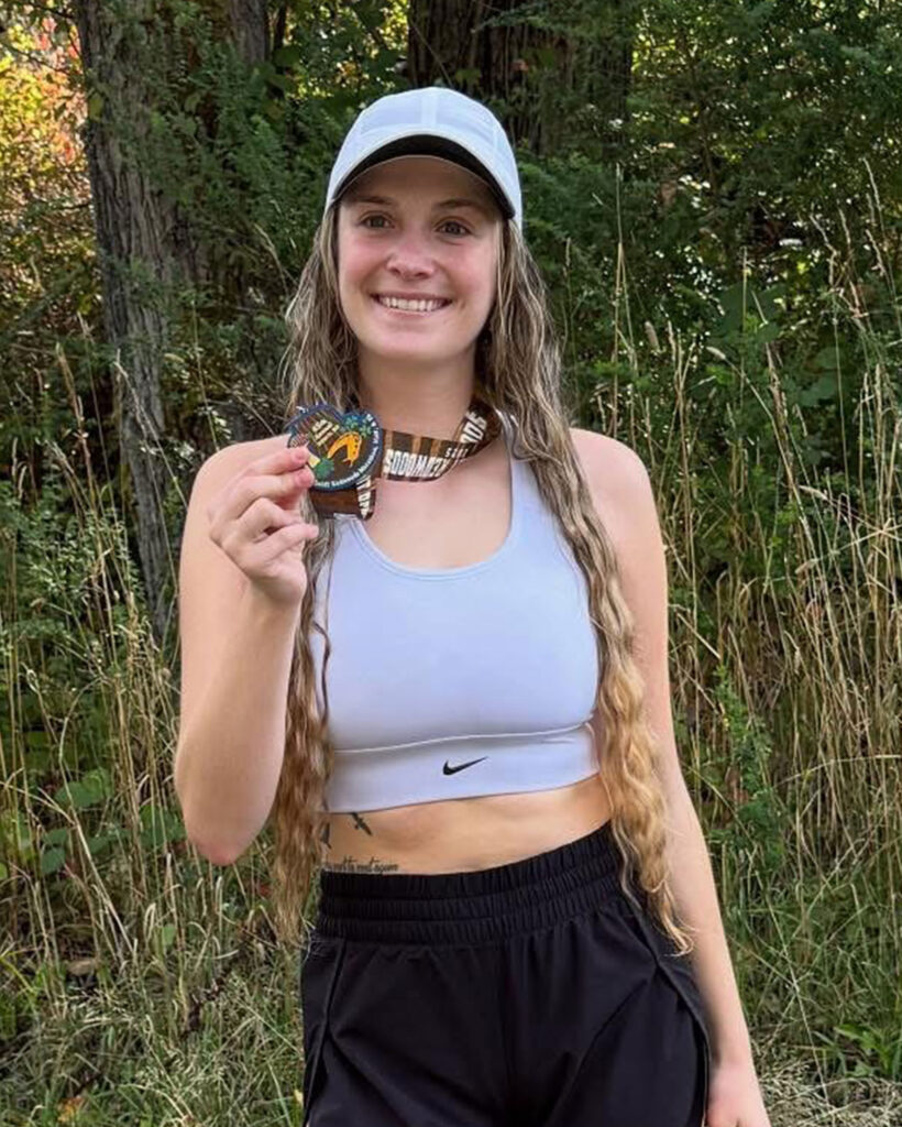 Athlete Rachelle standing on an outdoor running trail holding her medal from competing in a race. Rachelle is in fitness gear with a smile and a hat on.
